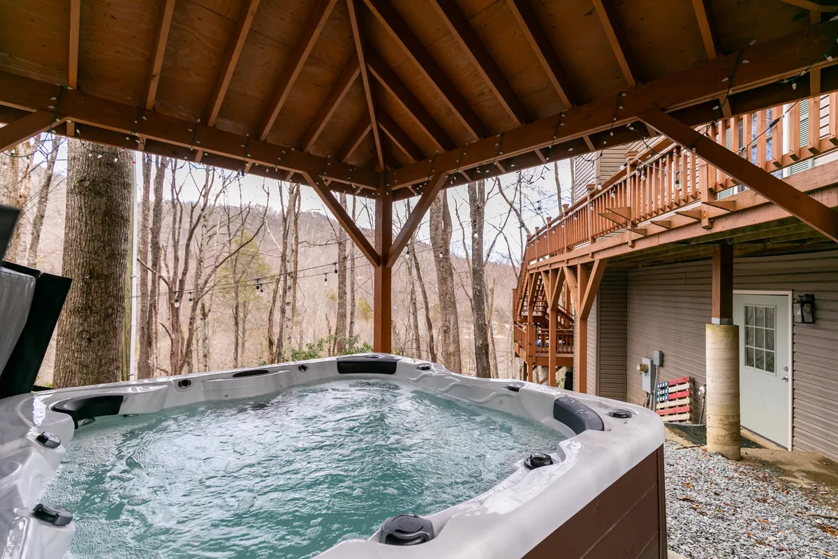 A serene exterior view featuring a bubbling hot tub set on a wooden deck, surrounded by lush trees. The inviting setup offers the perfect spot for stargazing or relaxing after a day of adventure.