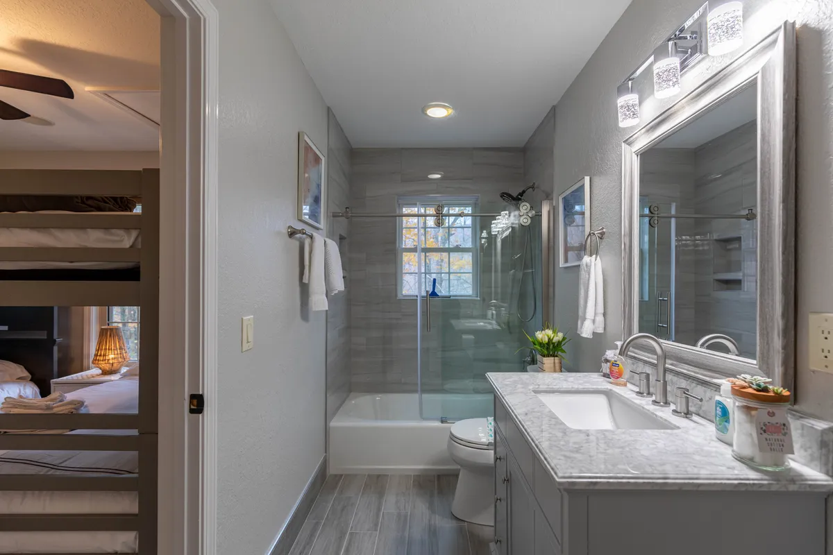 A functional and inviting bathroom that features a vanity with a sink and a glass shower enclosure, while a bunk bed is visible in the background. Sunlight fills the space, making it feel bright and practical for families or groups.