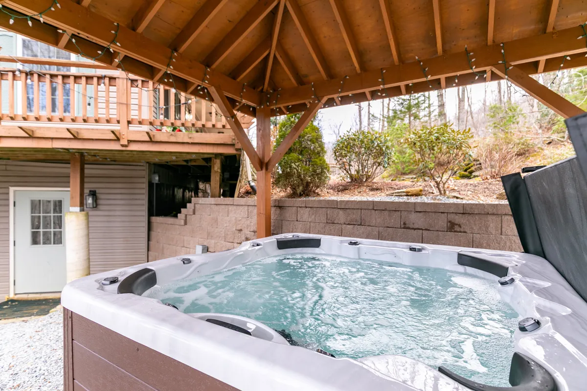 A tranquil outdoor hot tub nestled beneath a wooden structure, surrounded by lush greenery. The secluded atmosphere invites guests to unwind and socialize while soaking, with nature as a peaceful backdrop.