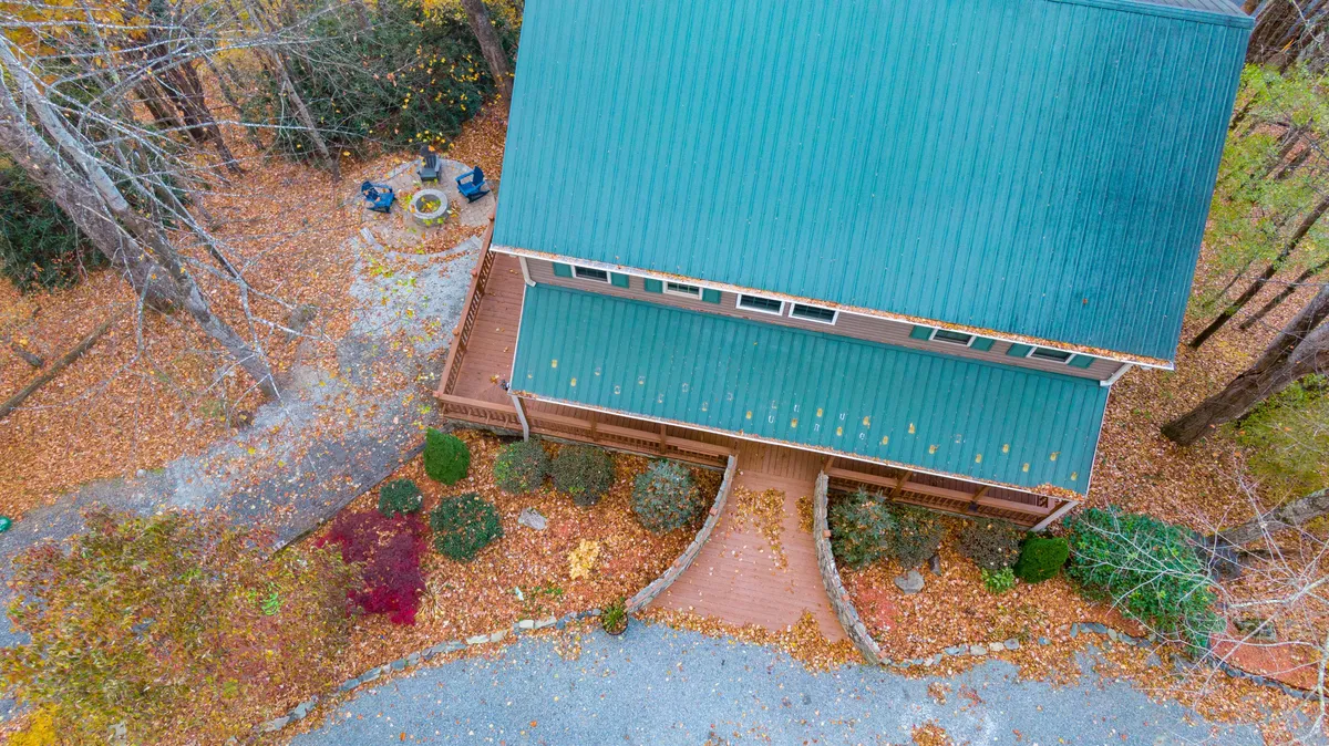An exterior view of the home showing a green metal roof, deck area, and a fire pit, all nestled among tall trees with autumn foliage. The setting highlights both the communal outdoor spaces and the peaceful wooded surroundings.