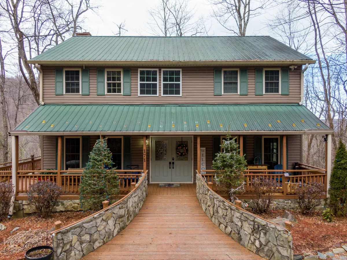The image showcases the inviting front porch of a single-family house with a distinctive green metal roof, a charming stone pathway leading up to the entrance, and a welcoming sign. The porch offers comfortable seating and overlooks lush greenery, hinting at serene sunset views and the perfect spot for gathering or relaxing.
