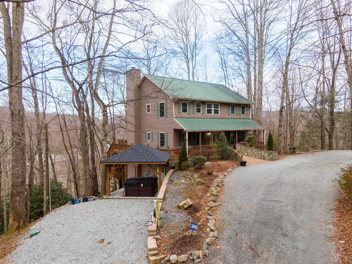 This image features a two-story house with a green metal roof nestled among mature trees, a spacious gravel driveway, and an inviting deck area. The natural surroundings convey a sense of peaceful seclusion, while the deck and yard provide ample space for outdoor gatherings and relaxation.