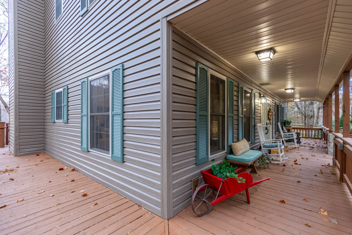 The image focuses on a wooden deck and porch adorned with green shutters, a red wheelbarrow, rocking chair, and decorative plants. The space feels comfortable and welcoming, ideal for relaxing, hosting gatherings, or enjoying a quiet moment while taking in seasonal views.
