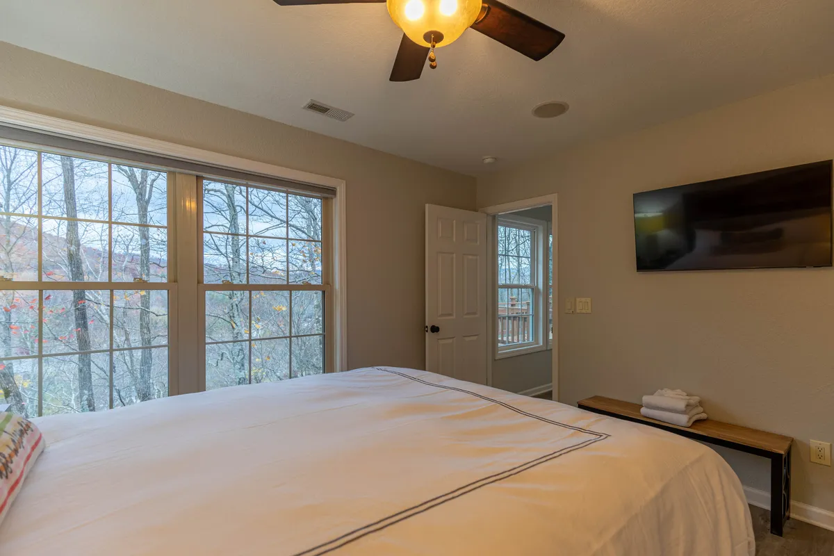 A spacious bedroom featuring a cozy bed, large windows with sunlight streaming in, a nightstand, and a wall-mounted television. The room feels welcoming and is equipped for both rest and entertainment.