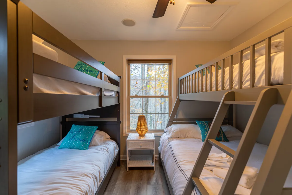 A playful bunk room with two sets of bunk beds, colorful pillows, and a decorative lamp. Natural light pours in through the window, creating a cheerful and inviting atmosphere ideal for kids or teens.