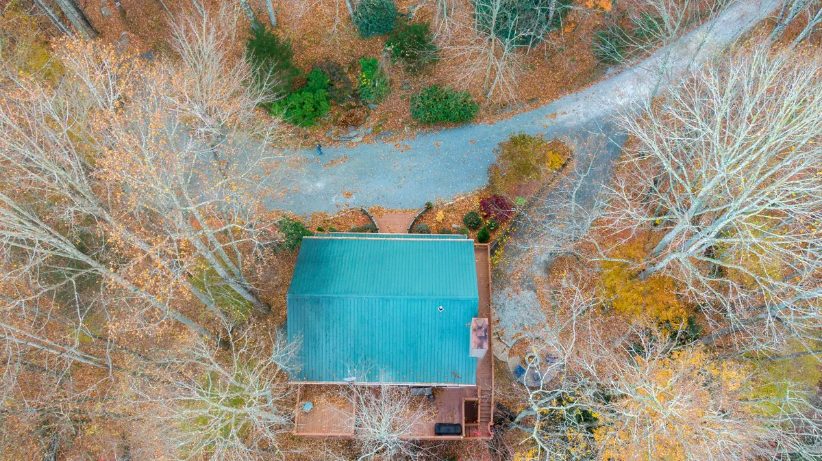 This wide-angle exterior shot features the house, the spacious driveway, and the surrounding forested landscape. The scene captures the appeal of the property as a base for nature walks, birdwatching, and enjoying the vivid changes in foliage across seasons.